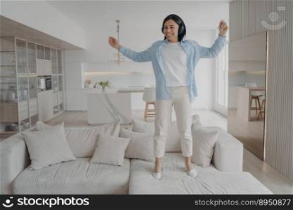 Excited young woman wearing wireless headphones jumping on sofa alone at home. Joyful energetic female having fun, enjoy musical sound, dancing listening to music on couch in living room on weekend.. Excited energetic woman wearing headphones having fun enjoying music jumping on sofa alone at home