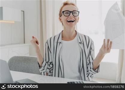 Excited young woman with paper bills in hands feeling euphoric, happy about finally paying down her mortgage debt, exclaiming yes with excitement while sitting at table with laptop. Excited young woman with paper bills in hands finally paying down her mortgage debt at home
