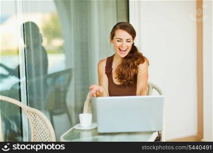 Excited young woman working on laptop on terrace with cup of coffee