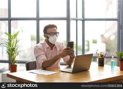 Executive in a mask texting on his phone while working on his laptop