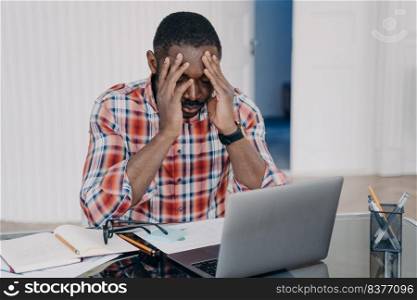 Exhausted african american man suffer headache after working at laptop. Tired frustrated black guy holds head by hands feel migraine pain at workplace. Stress at work, occupational burnout.. Exhausted african american man suffer headache at laptop. Stress at work, occupational burnout