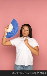 Exhausted african american woman with closed eyes using blue fan suffer from heat sweating, feels sluggish. Black female cooling in hot summer weather, high temperature, isolated on pink background.. Exhausted african woman using paper fan suffer from heat sweating, feels sluggish isolated on pink.