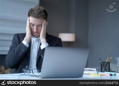 Exhausted man German legal consultant sits at workplace at home office in formal suit, took off glasses, gently touches his temples with fingers, feels headache while working in online court session. Exhausted legal consultant sitting at home office gently touches his temples with fingers