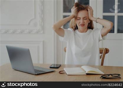 Exhausted woman holds her head. Freelancer has deadline. Stressed mid adult businesswoman is working from home in evening. Tired european lady is manager sitting at the desk at laptop.. Exhausted woman holds her head. Freelancer has deadline and working from home in evening.