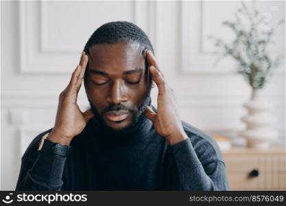 Exhausted young Afro American male entrepreneur home office worker sitting with eyes closed and with hands on his head slowly and gently massaging temples with fingers, consequences of overwork. Exhausted young Afro American male entrepreneur sitting with eyes closed and with hands on his head