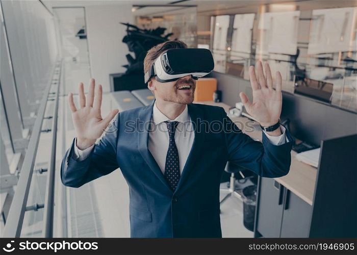 Exited businessman stands in office interior after finishing his workday in virtual reality VR headset while trying to touch with hands virtual objects in digital simulation during gaming experience. Exited businessman stands in office interior after finishing workday in virtual reality VR headset