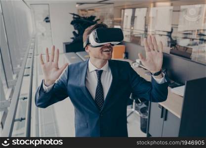 Exited businessman stands in office interior after finishing his workday in virtual reality VR headset while trying to touch with hands virtual objects in digital simulation during gaming experience. Exited businessman stands in office interior after finishing workday in virtual reality VR headset