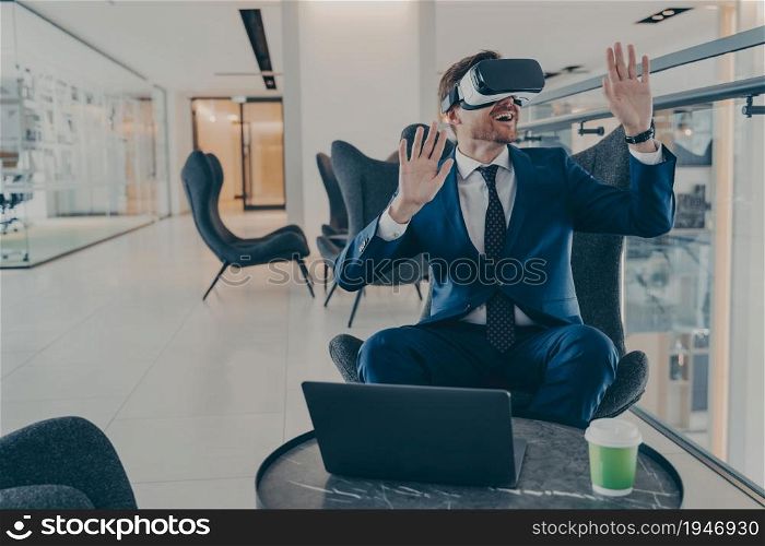 Exited CEO in VR headset enthusiastically testing app on laptop sitting in business centre lobby. Office worker immersed in virtual reality, innovative method of managing business projects in 3D touch. Exited CEO in VR headset enthusiastically testing app on laptop sitting in business centre lobby