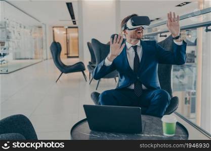 Exited CEO in VR headset enthusiastically testing app on laptop sitting in business centre lobby. Office worker immersed in virtual reality, innovative method of managing business projects in 3D touch. Exited CEO in VR headset enthusiastically testing app on laptop sitting in business centre lobby