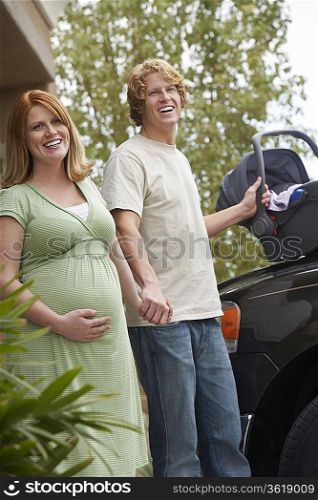 Expectant couple with baby carrier by car, portrait