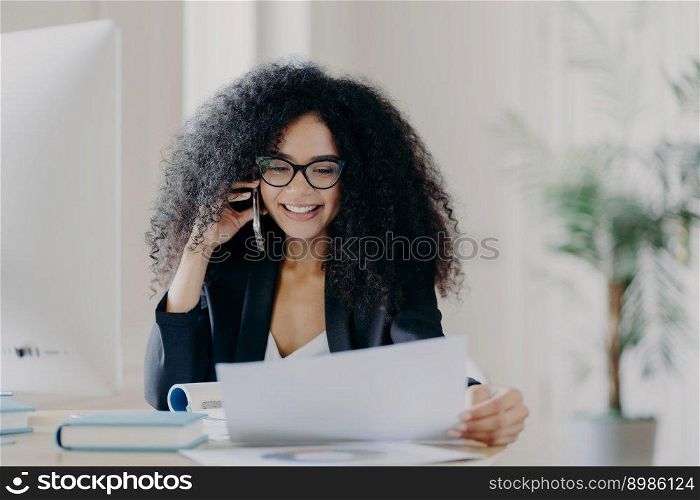 Experienced female CEO has telephone conversation, solves problem, focused in documents, wears spectacles and formal wear, poses at work place, has cheerful expression. Administrative manager