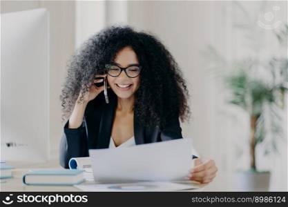 Experienced female CEO has telephone conversation, solves problem, focused in documents, wears spectacles and formal wear, poses at work place, has cheerful expression. Administrative manager