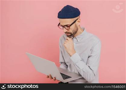 Experienced male freelancer develops new strategy, holds modern laptop computer, keeps hand on chin, wears formal clothing, busy with distant work, isolated over pink background, copy space.