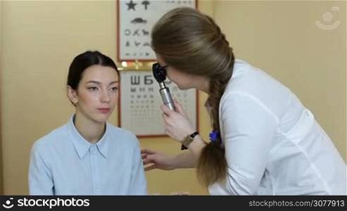 Experienced optometrist examining attractive female patient through ophthalmoscope in ophthalmology clinic. Ophthalmologist with optical device checking patient&acute;s eyesight using ophthalmoscope in oculist office. Ophthalmology and eye care concept.