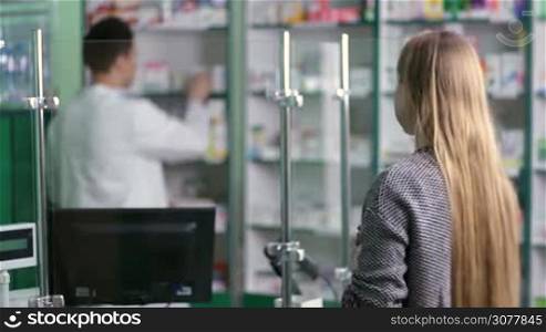 Experienced pharmacist in white coat counseling female customer in modern pharmacy, view of the pharmacist over the client&acute;s shoulder. Handsome friendly drugist with stethoscope selling medicine at the drugstore. Health care and pharmacology concept