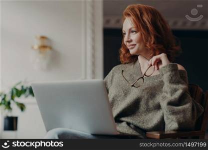 Experienced redhead female entrepreneur makes plans, looks aside with dreamy expression, uses modern technology, holds spectacles, wears casual jumper, enjoys distance education and e learning