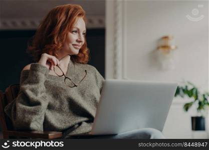 Experienced redhead female entrepreneur makes plans, looks aside with dreamy expression, uses modern technology, holds spectacles, wears casual jumper, enjoys distance education and e learning