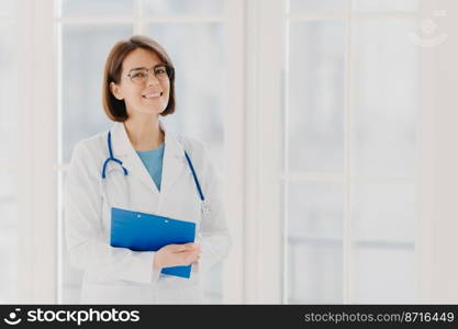 Experienced woman pediatrician stands with clipboard in cabinet, wears white medical coat with stethoscope, gives excellent medical treatment, has happy expression, ready to give consultancy