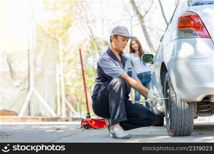 Expertise mechanic man in uniform using force trying to unscrew the wheel bolts nuts and help a woman for changing car wheel on the highway, car service, repair, maintenance concept.