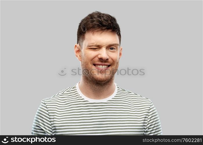 expression and people concept - smiling young man in striped t-shirt winking over grey background. smiling young man in striped t-shirt winking