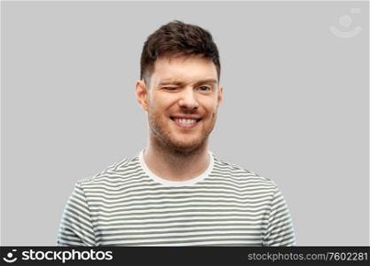 expression and people concept - smiling young man in striped t-shirt winking over grey background. smiling young man in striped t-shirt winking