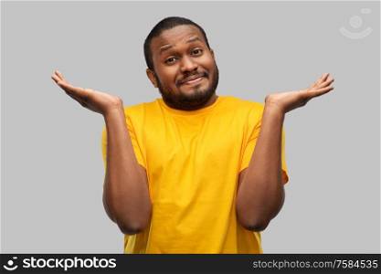 expressions and people concept - clueless young african american man in yellow t-shirt shrugging over grey background. african american man in yellow t-shirt shrugging