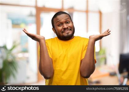 expressions and people concept - clueless young african american man in yellow t-shirt shrugging over office background. african american man in yellow t-shirt shrugging