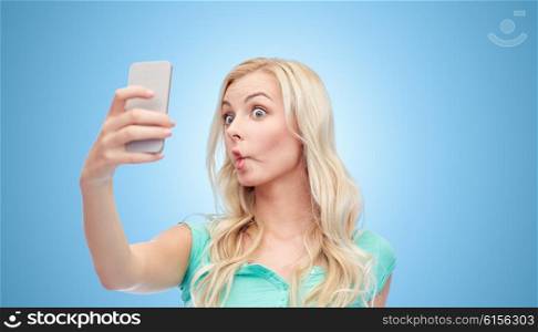 expressions, technology and people concept - funny young woman or teenage girl taking selfie with smartphone and making fish face over blue background
