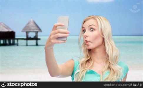 expressions, technology, travel, tourism and people concept - funny young woman or teenage girl taking selfie with smartphone and making fish face over tropical beach with bungalow background