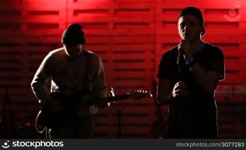 Expressive teenage singer with eyes closed holding microphone and singing song soulfully with stylish guitarist playing on electric guitar at the background. Rock group performing at concert on illuminated stage.