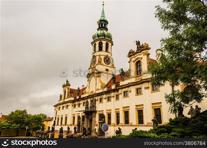 Exterior facade of Loreta church in Prague: green rooftop on white belfry, white walls, red rooftop. In a cloudy gloomy day, green trees park