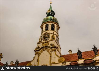 Exterior facade of Loreta church in Prague: green rooftop on white belfry, white walls, red rooftop. In a cloudy gloomy day, green trees park