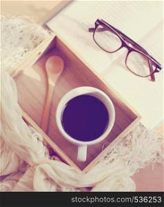 Eyeglasses and book with black coffee on wooden tray, retro filter effect