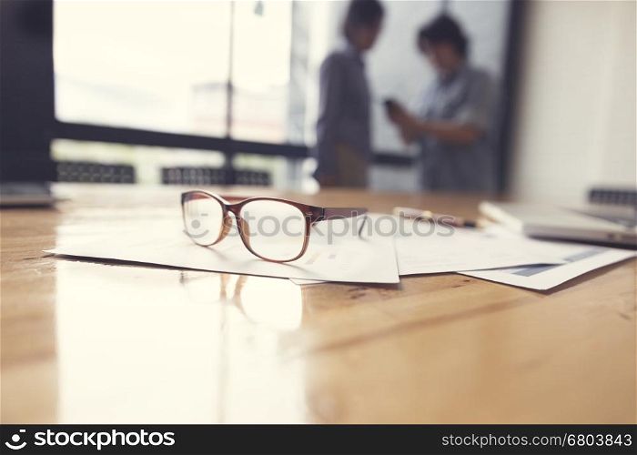eyeglasses, business document and laptop computer notebook on wooden table with people background, selective focus and vintage tone