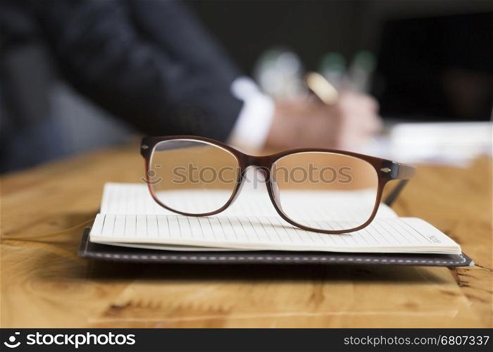 eyeglasses on notebook with background of businessman in suit working with document on office desk