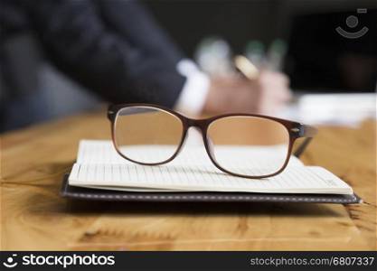 eyeglasses on notebook with background of businessman in suit working with document on office desk