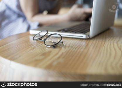 eyeglasses with laptop computer notebook on wooden desk with background of woman sitting on chair