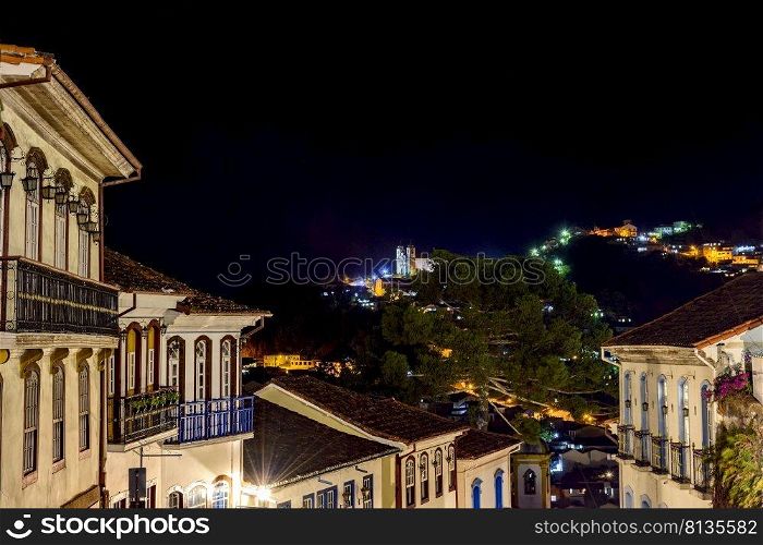 Facades of houses in colonial architecture on an old cobblestone street in the city of Ouro Preto illuminated at night with historic church in background. Facades of houses in colonial architecture on an old cobblestone street illuminated at night with historic church in background