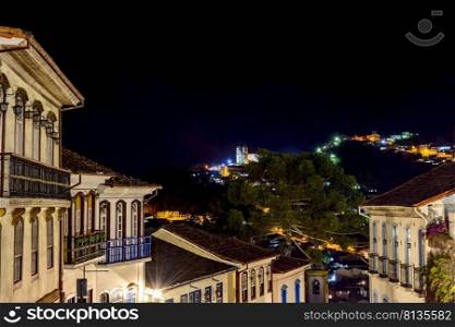 Facades of houses in colonial architecture on an old cobblestone street in the city of Ouro Preto illuminated at night with historic church in background. Facades of houses in colonial architecture on an old cobblestone street illuminated at night with historic church in background