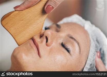 Face Massage with Wooden Gua Sha Plate Shape Massager. Skin Care treatment at beauty spa center. Face Massage with Wooden Gua Sha Plate Shape Massager