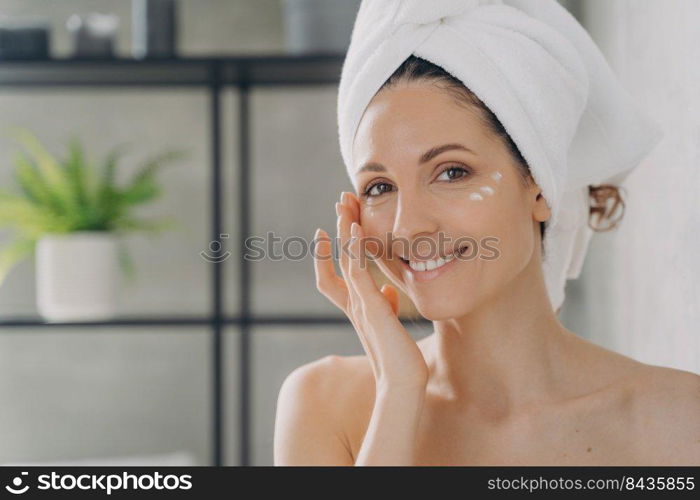 Face of beautiful european lady which applies cream. Attractive feminine caucasian girl wrapped in towel after bathing and hair washing. Happy young woman takes shower at home. Beauty routine.. Face of beautiful european lady which applies cream. Attractive feminine girl beauty routine.