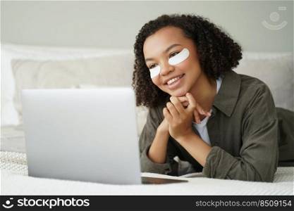 Face of carefree african american girl which applies eye patches and relaxing at home watching video on laptop. Attractive curly girl is grooming herself in bedroom and having fun. Beauty routine.. Face of carefree african american girl which applies eye patches and watching video on laptop.