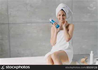 Face toner or micellar water advertising concept. Clean skin of beautiful caucasian woman wrapped in towel after bathing. Young happy woman takes shower at home holding face washing foam in bottle.. Young happy woman takes shower at home. Girl is holding face washing foam in bottle.