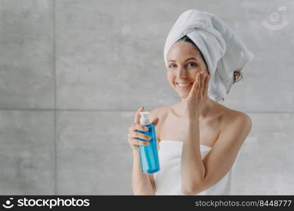 Face toner or micellar water advertising concept. Happy young woman touches pure skin. Gorgeous caucasian woman wrapped in towel after bathing applying toner in flacon. Face washing foam in bottle.. Face toner or micellar water. Gorgeous caucasian woman applying toner in flacon touching pure skin.