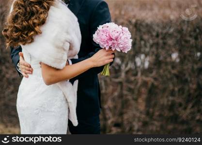 Faceless bride and groom in festive clothes, embrace and stand closely, woman in white dress holds wedding bouquet, stand outside. Newlyweds. Special occasion. Romantic relationship concept.