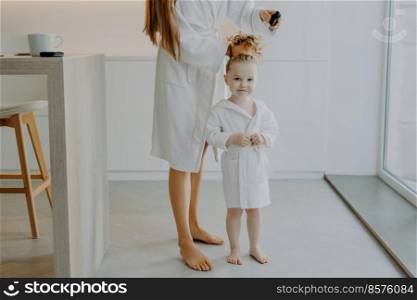 Faceless mother makes pony tail from daughters curly hair uses comb. Cute little three year old girl in white bathrobe after taking shower poses near mom at home. Children parenthood beauty time