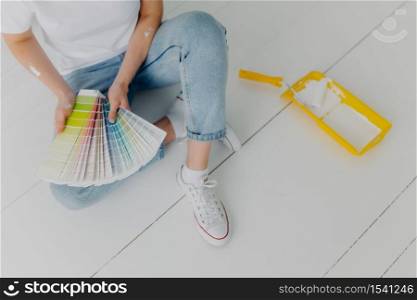 Faceless woman in jeans holds color samples, chooes best tone for refurbishment walls, sits on white floor, uses tray and paint roller for house remodeling. Home makeover and renovation concept
