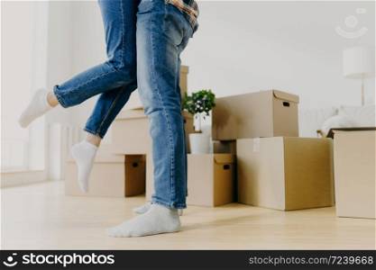 Faceless young couple move in new home, man lifts woman, have fun, surrounded with unpacked cardboard boxes, start new life in recently bought abode. Family, relocation and moving day concept
