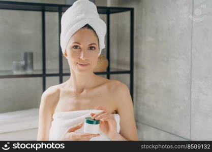 Facial cream advertising banner. European lady applies face cream. Attractive girl wrapped in towel after bathing and hair washing is holding cream jar. Happy woman takes shower at home.. Facial cream advertising banner. Girl wrapped in towel after bathing and hair washing holding jar.