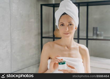 Facial cream advertising banner. European lady applies face cream. Attractive girl wrapped in towel after bathing and hair washing is holding cream jar. Happy woman takes shower at home.. Facial cream advertising banner. Girl wrapped in towel after bathing and hair washing holding jar.